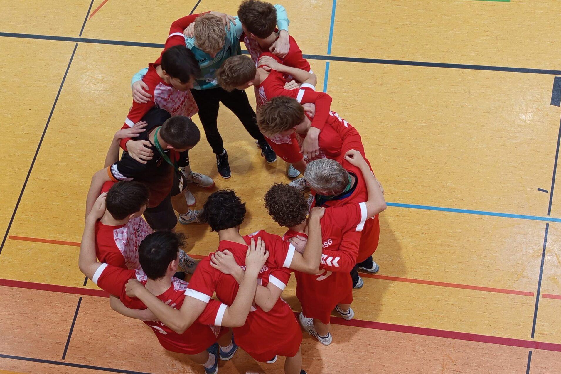 Gruppe von Sportlern in roten Trikots bildet einen Kreis und umarmt sich auf einem hellen Sportboden.