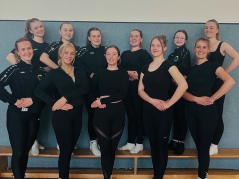 Gruppe von zehn Frauen in Sportkleidung, posierend auf einer Holzbank in einer Turnhalle.