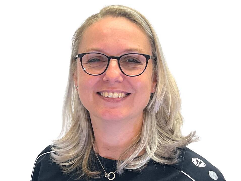 Portrait einer lächelnden Frau mit blonden, langen Haaren, Brille und schwarzem Sportshirt mit Logo.