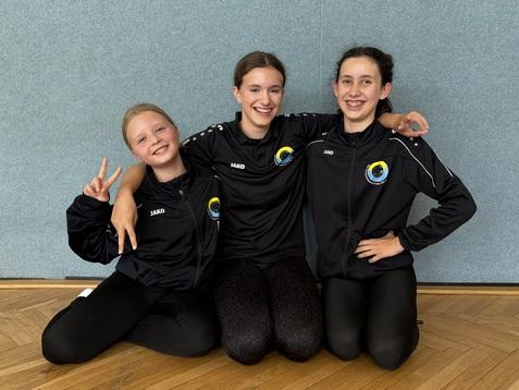 Drei Mädchen im Sportanzug sitzen lächelnd auf dem Boden, arm in arm, vor einer grauen Wand.