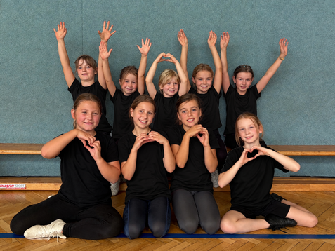 Zehn Kinder in schwarzen T-Shirts posieren l&auml;chelnd in einer Sporthalle, einige formen mit den H&auml;nden Herzen.