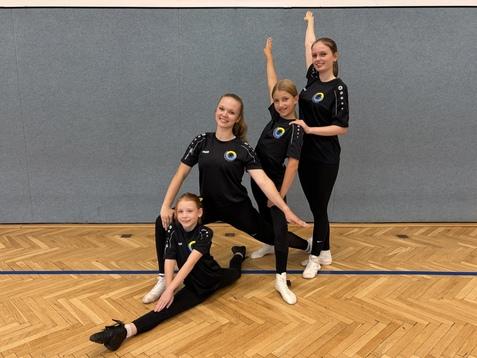 Vier Tänzerinnen in schwarzen Outfits posieren in einer Sporthalle auf einem Parkettboden.