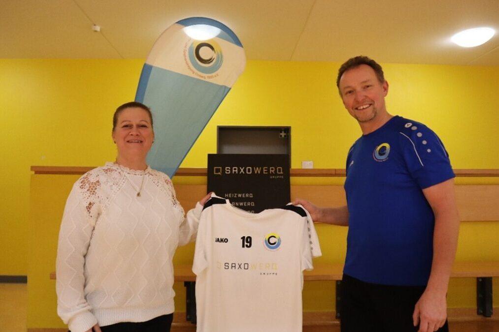 Zwei Personen präsentieren ein Sporttrikot vor einer gelben Wand, mit einem aufgestellten Banner im Hintergrund.