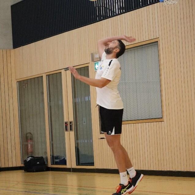 Ein Volleyballspieler springt zum Aufschlag in einer Sporthalle mit Holzverkleidung. Der Ball befindet sich in der Luft.