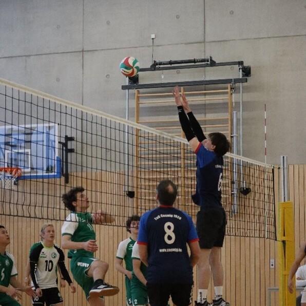 Zwei Volleyballspieler springen am Netz, während ein dritter Spieler den Ball angreift. Sporthalle mit Holzwänden im Hintergrund.