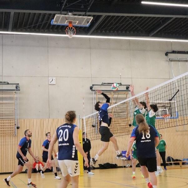 Spieler in blauen Trikots springen, um einen Volleyball über das Netz zu schlagen; grüne Mannschaft blockt. Halle mit Holzwand.