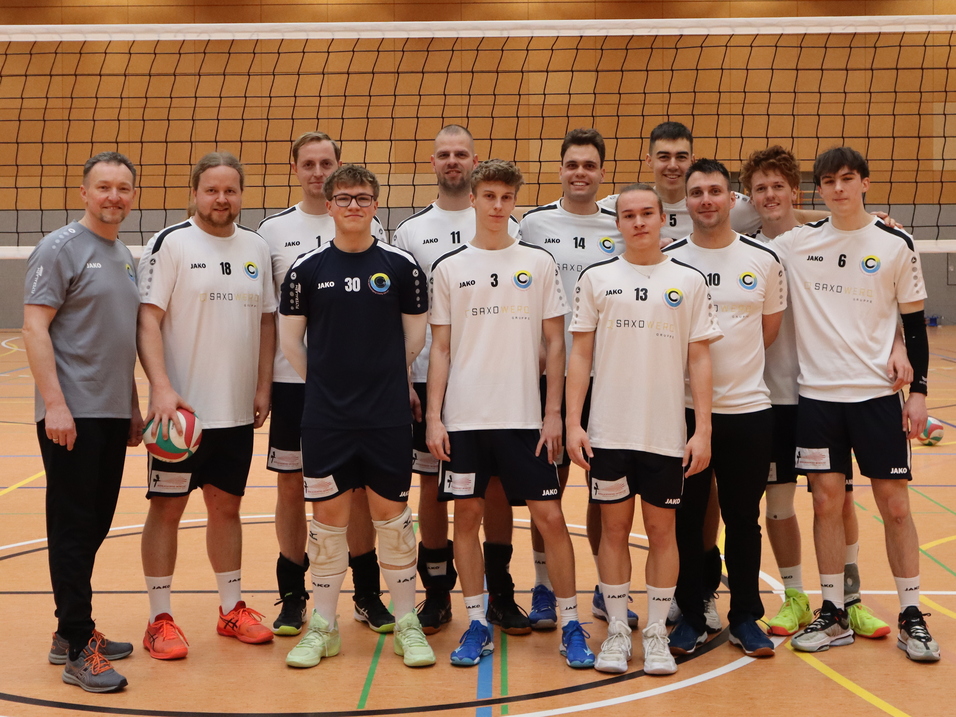 Gruppenfoto eines Volleyballteams in Sportkleidung, stehend vor dem Netz in einer Halle.