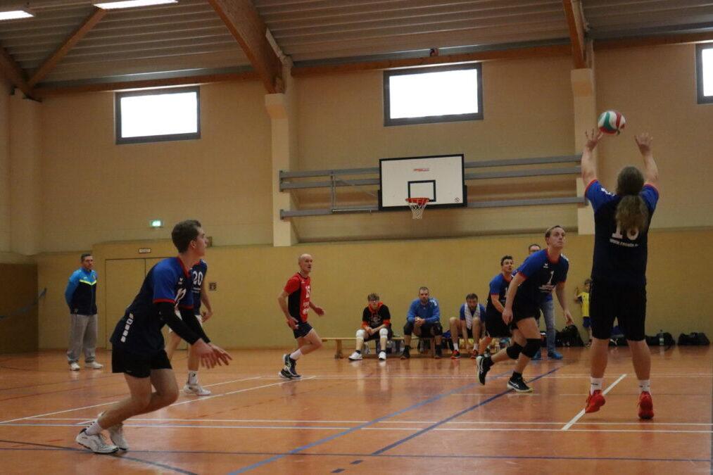 Volleyballspiel in einer Halle, ein Spieler passt den Ball, während andere agieren. Zuschauer sitzen im Hintergrund.