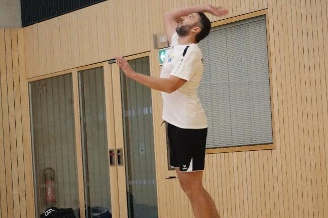 Ein Volleyballspieler springt zum Aufschlag in einer Sporthalle mit Holzverkleidung. Der Ball befindet sich in der Luft.