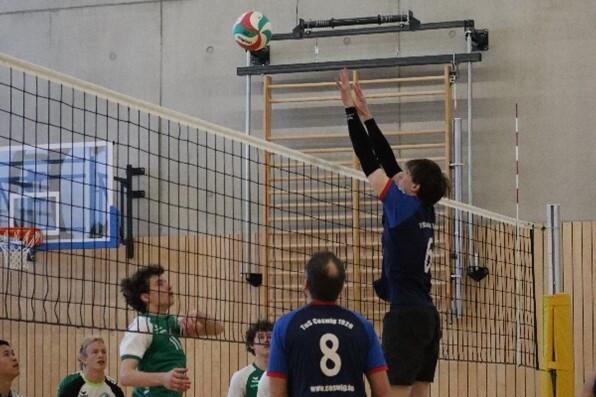 Zwei Volleyballspieler springen am Netz, während ein dritter Spieler den Ball angreift. Sporthalle mit Holzwänden im Hintergrund.