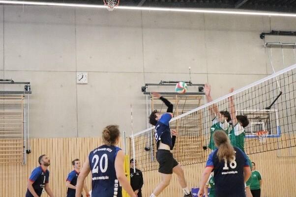 Spieler in blauen Trikots springen, um einen Volleyball über das Netz zu schlagen; grüne Mannschaft blockt. Halle mit Holzwand.