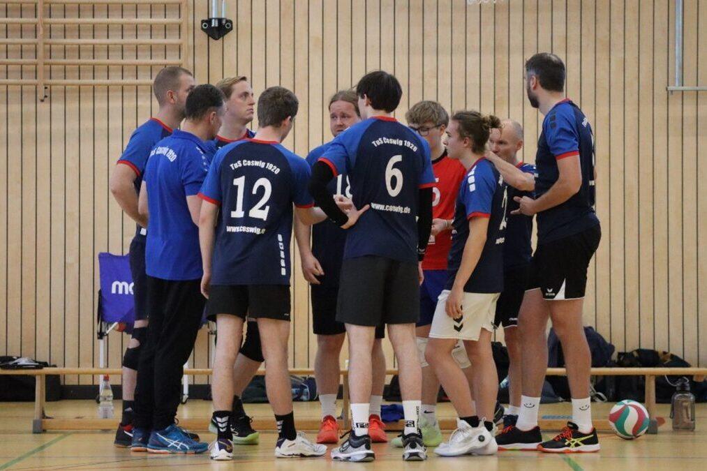 Eine Gruppe von Volleyballspielern in blauen Trikots steht im Kreis, besprechend auf einem Hallenboden mit Holzverkleidung.