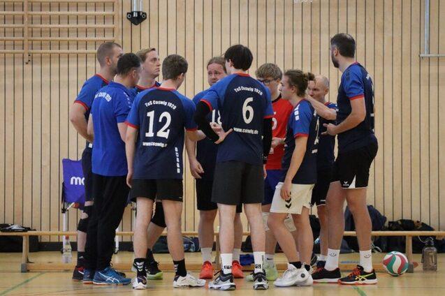Eine Gruppe von Volleyballspielern in blauen Trikots steht im Kreis, besprechend auf einem Hallenboden mit Holzverkleidung.
