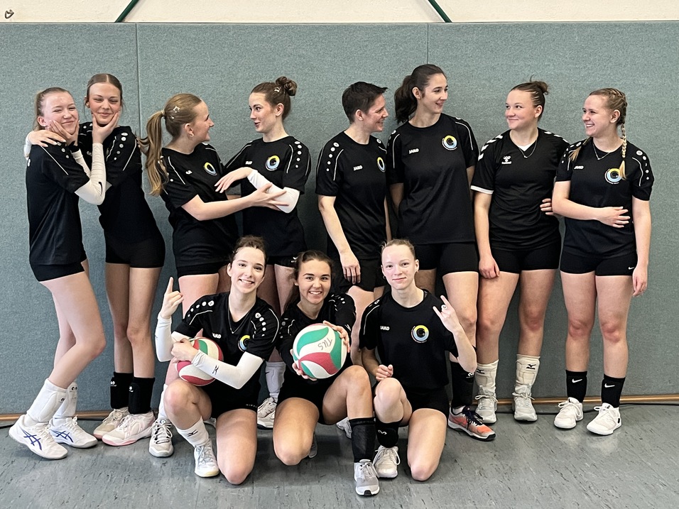 Gruppe von elf M&auml;dchen in Sportkleidung, l&auml;chelnd, mit Volleyb&auml;llen, in einer Sporthalle vor grauer Wand.