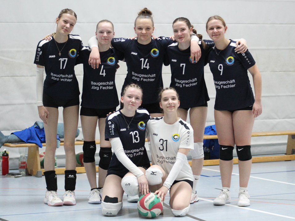 Gruppe von sieben Volleyballspielerinnen in schwarzen Trikots, l&auml;chelnd auf einem Sportplatz zusammengestanden.