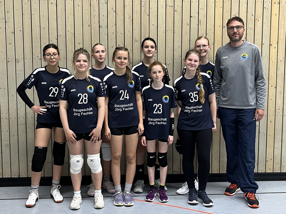 Mannschaftsbild von acht Volleyballspielerinnen in einheitlichen Trikots und einem Trainer vor einer Holzwand.