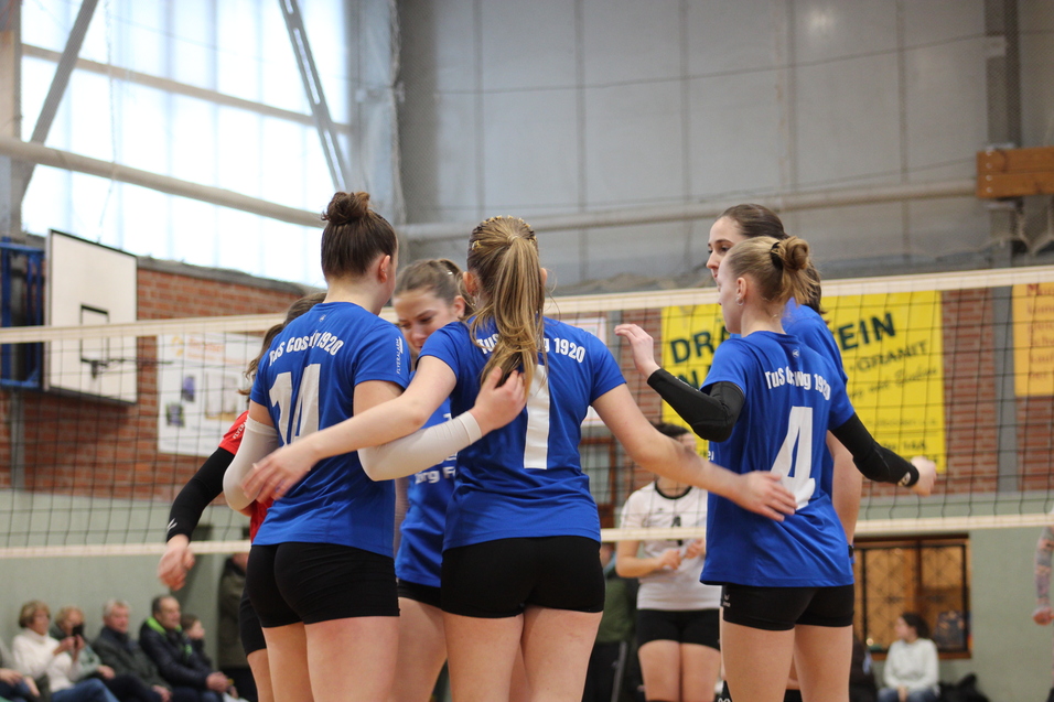 Sechs weibliche Volleyballspieler in blauen Trikots umarmen sich vor dem Netz in einer Sporthalle.