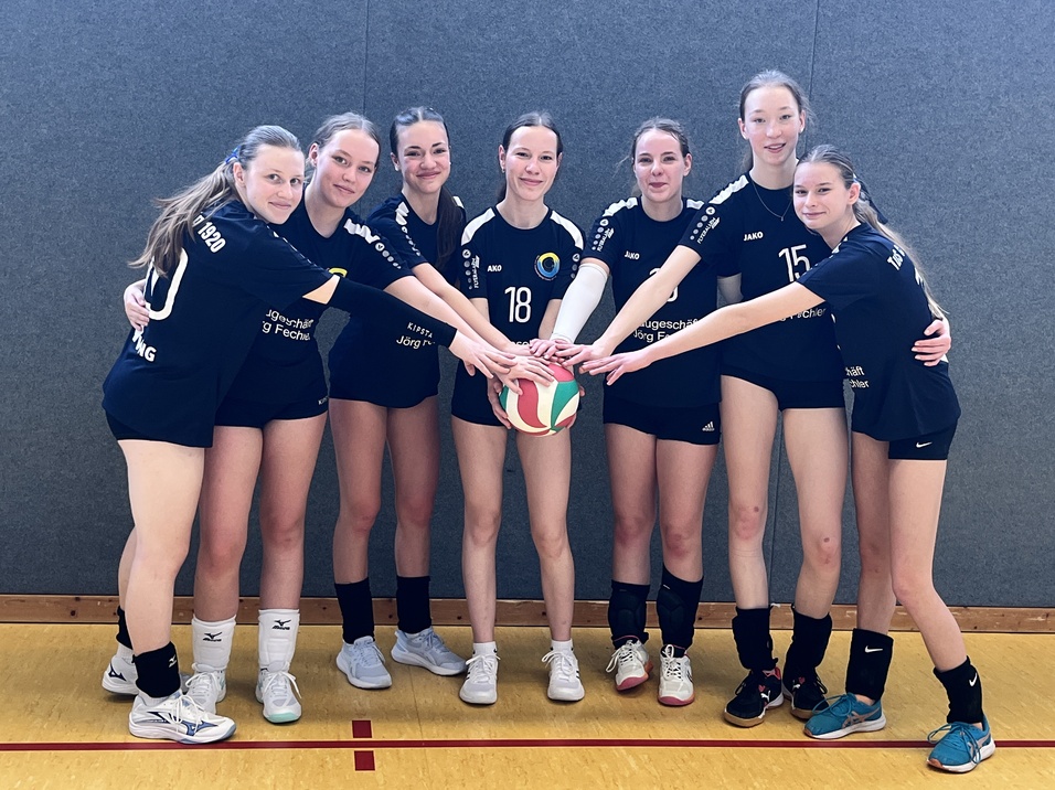 Gruppe von acht Volleyballspielerinnen in schwarzen Trikots um einen Volleyball versammelt auf einem Sporthallboden.