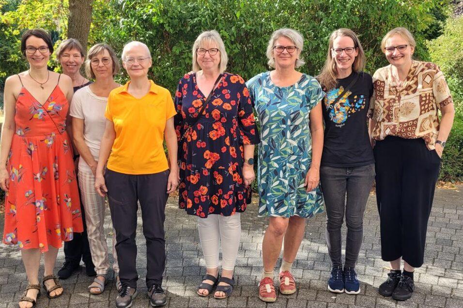 Gruppe von acht Frauen steht lächelnd nebeneinander in einem grünen Garten, modebewusst gekleidet und freundlich.