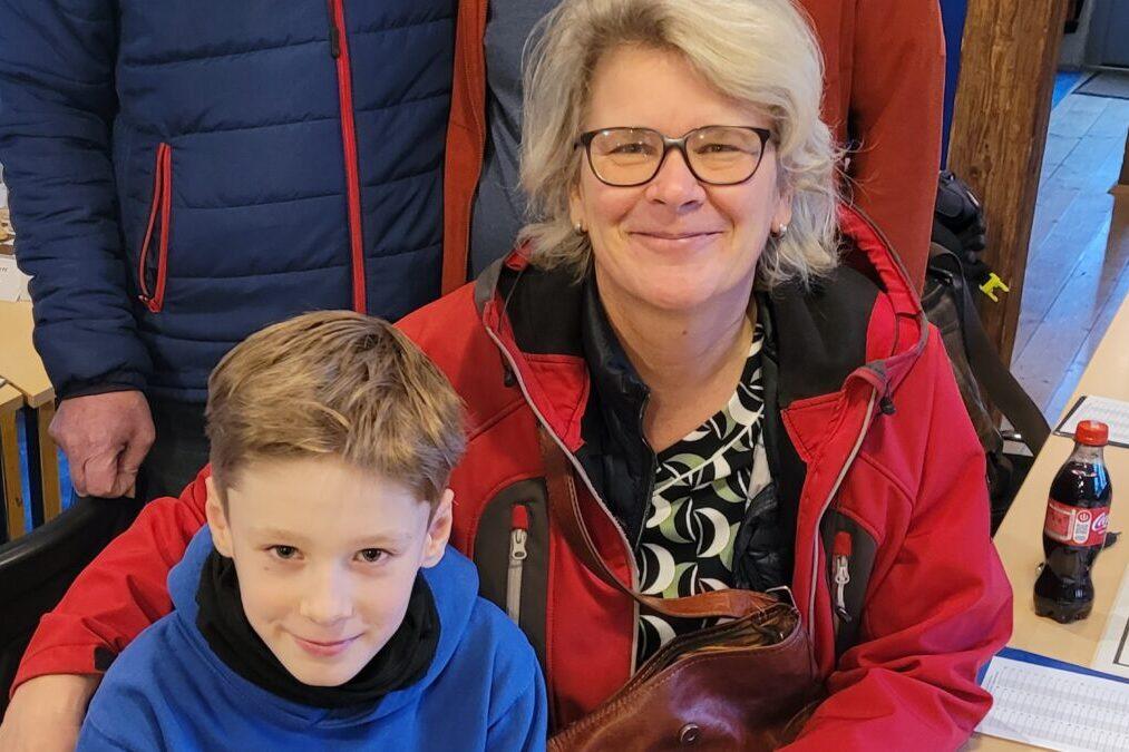 Eine Frau mit Brille und roter Jacke sitzt neben einem Jungen in blauer Jacke, beide lächeln in die Kamera.