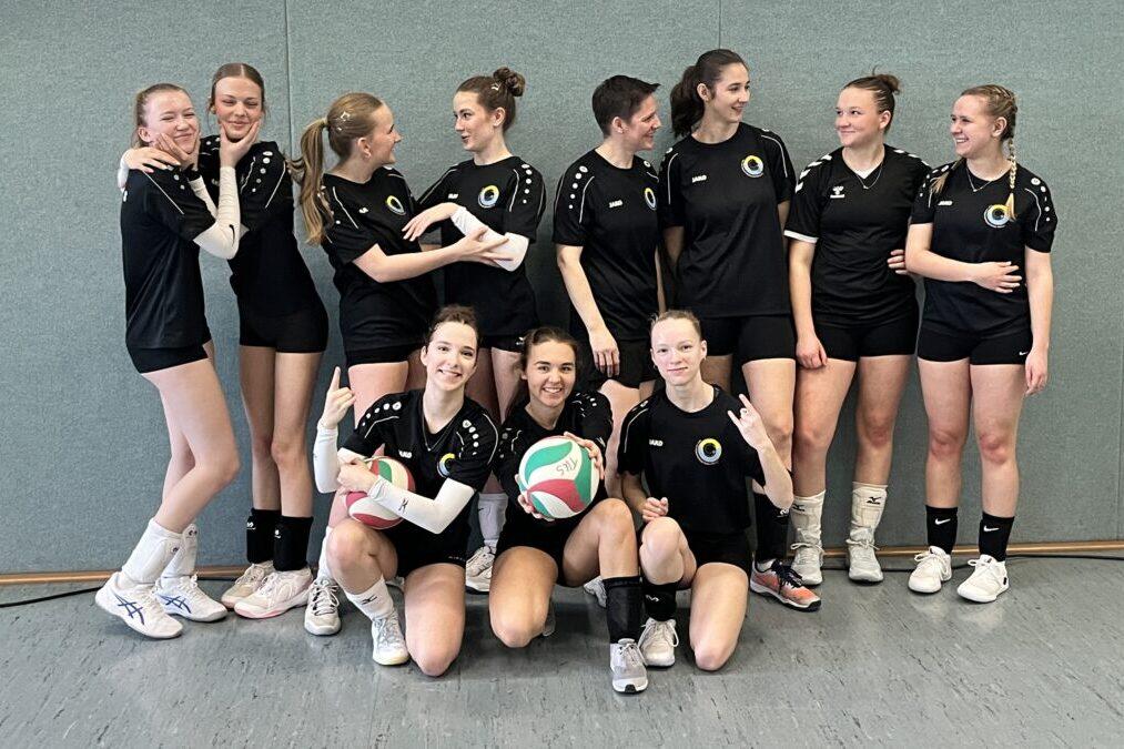 Gruppe von Volleyballspielerinnen in schwarzen Trikots posiert lächelnd, einige halten einen Volleyball in der Hand.