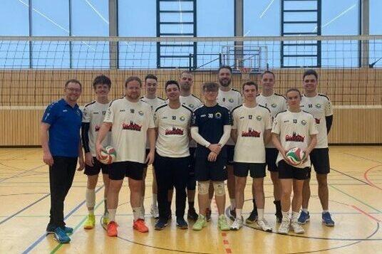Gruppe von zehn Volleyballspielern in Sportkleidung, stehen zusammen in einer Sporthalle vor einem Volleyballnetz.