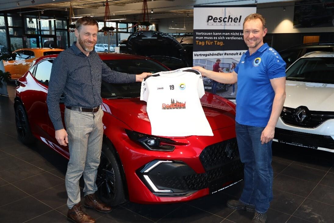 Zwei Männer präsentieren ein Trikot vor einem roten Sportwagen in einem Autohaus.