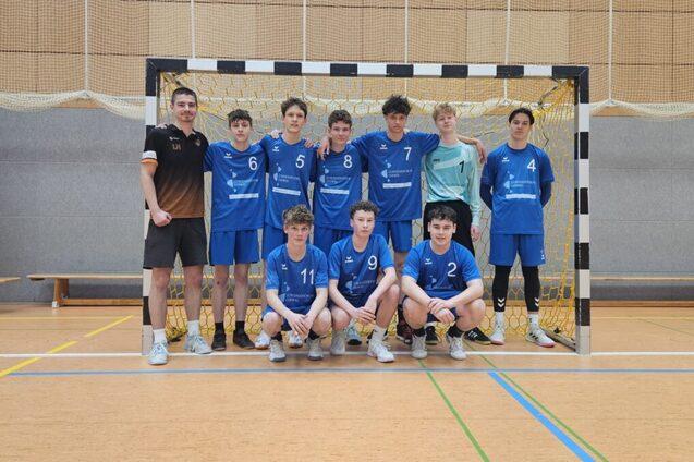 Gruppe von zehn Handballspielern in blauen Trikots steht vor einem Tor in einer Sporthalle.