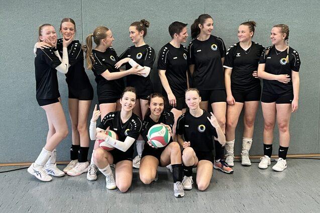 Gruppe von Volleyballspielerinnen in schwarzen Trikots posiert l&auml;chelnd, einige halten einen Volleyball in der Hand.