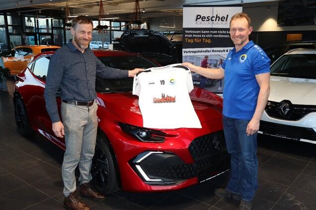 Zwei M&auml;nner pr&auml;sentieren ein Trikot vor einem roten Sportwagen in einem Autohaus.
