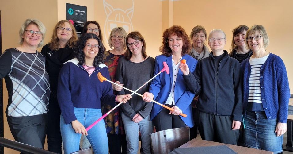 Gruppe von zwölf Frauen in einem Raum, die mit Tischtennisschlägern und Bällen posieren.