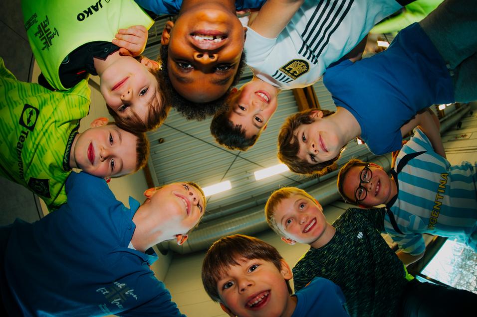 Gruppe von acht lächelnden Kindern in Sportswear, die sich in einem Kreis um eine Kamera versammeln.