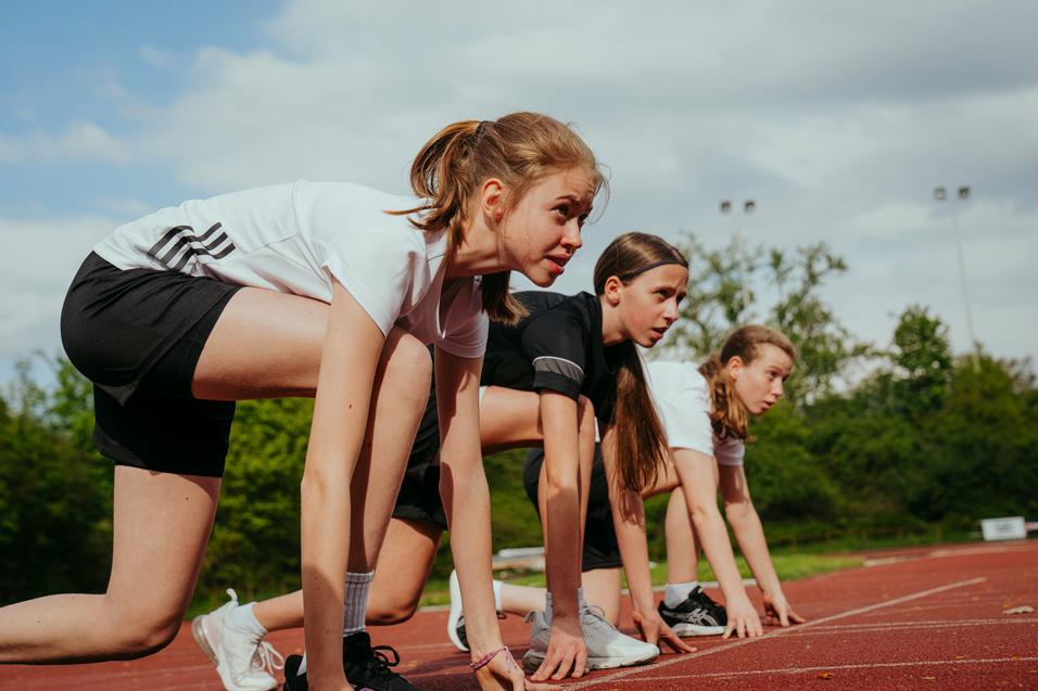 Drei junge Athletinnen in sportlicher Kleidung stehen in Startposition auf einer Laufbahn.