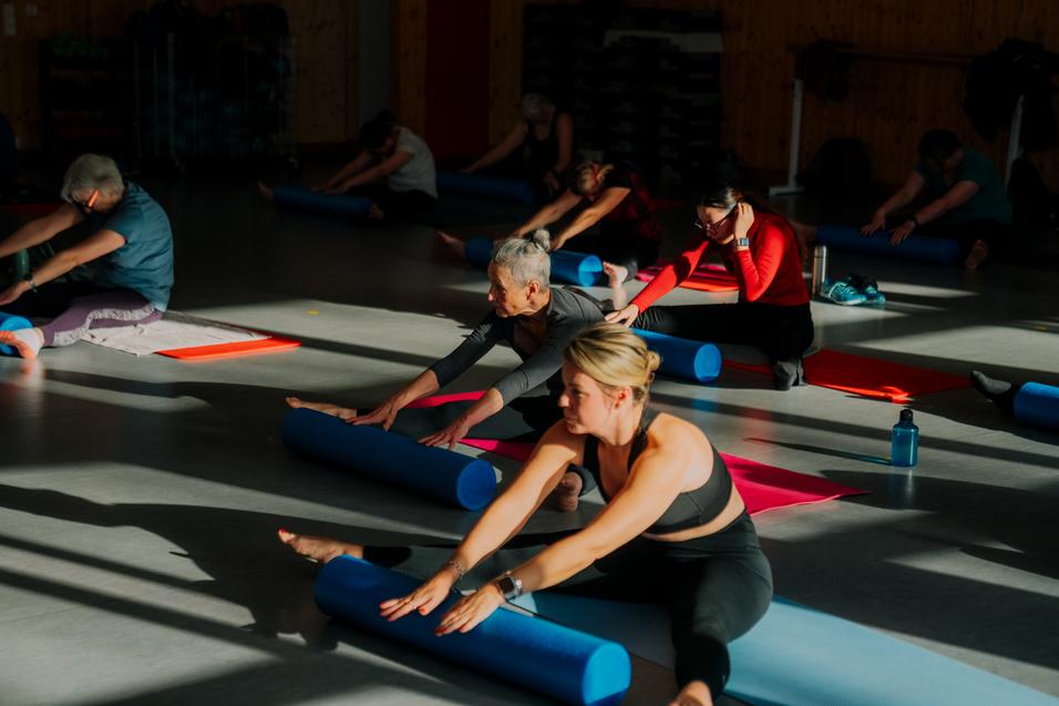 Gruppe von Frauen in Sportkleidung dehnt sich auf Yogamatten mit blauen Schaumstoffrollen in einem gut beleuchteten Raum.