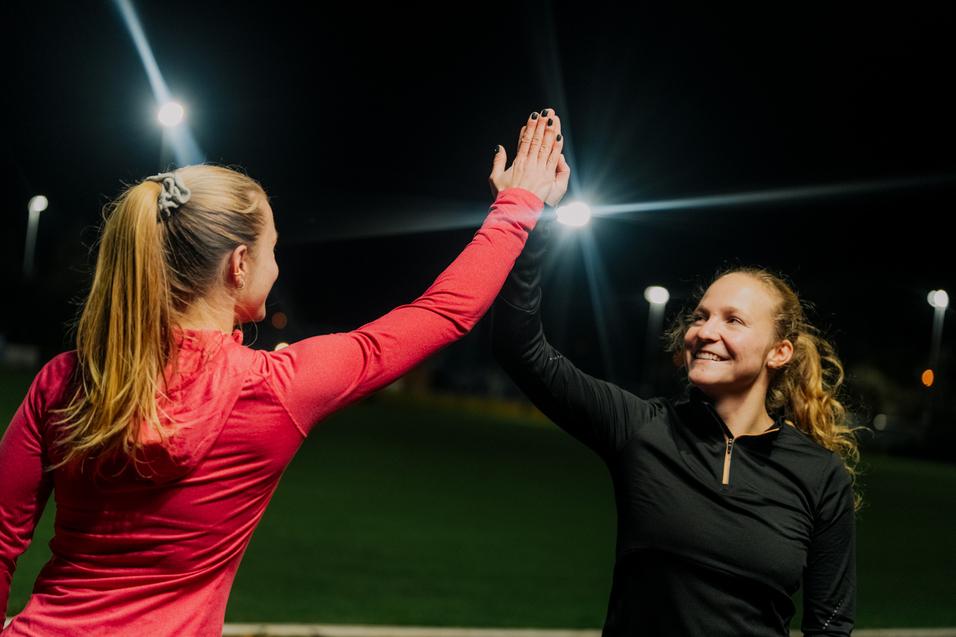 Zwei Frauen hintereinander geben sich bei Nacht einen High-Five, beleuchtet von Scheinwerfern auf einem Sportfeld.
