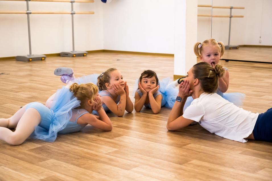 Fünf kleine Balletttänzerinnen in blauen Tüllkleidern liegen lächelnd auf dem Boden und betrachten eine Trainerin.