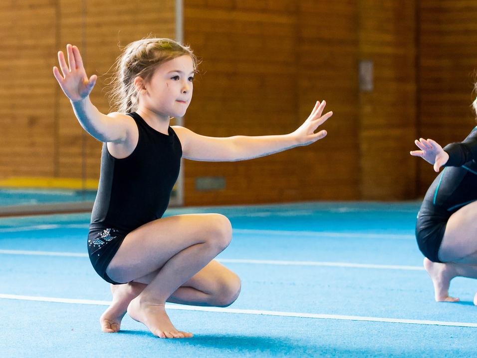 Zwei Mädchen in Gymnastikanzügen üben auf einer blauen Matte, eine kniet mit ausgestreckten Armen.