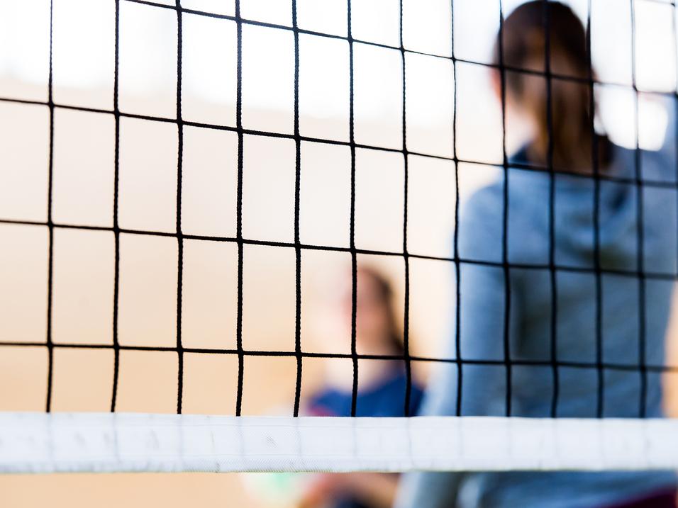 Nahaufnahme eines Volleyballnetzes mit unscharfen Spielern im Hintergrund in einer Sporthalle.